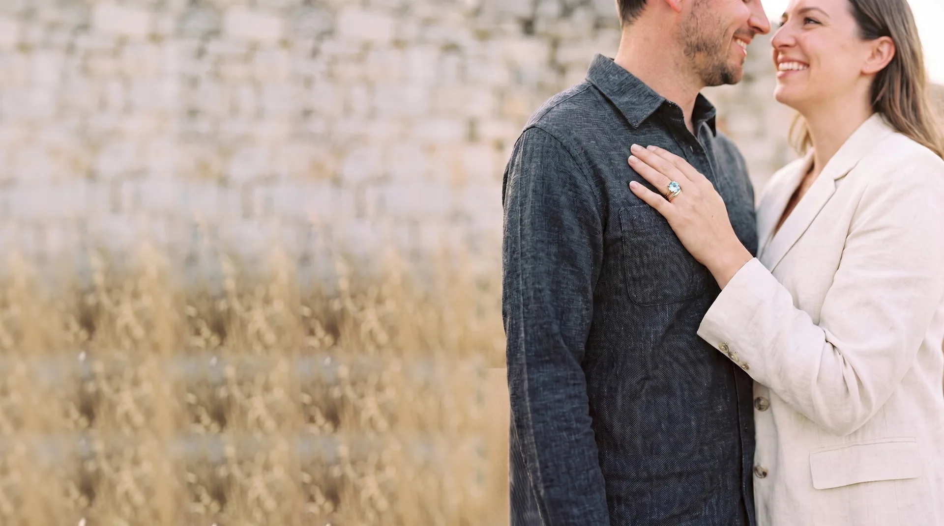 A couple outdoors in evening light, her left hand on his chest wearing a pale blue solitaire engagement ring and a slim gold wedding band